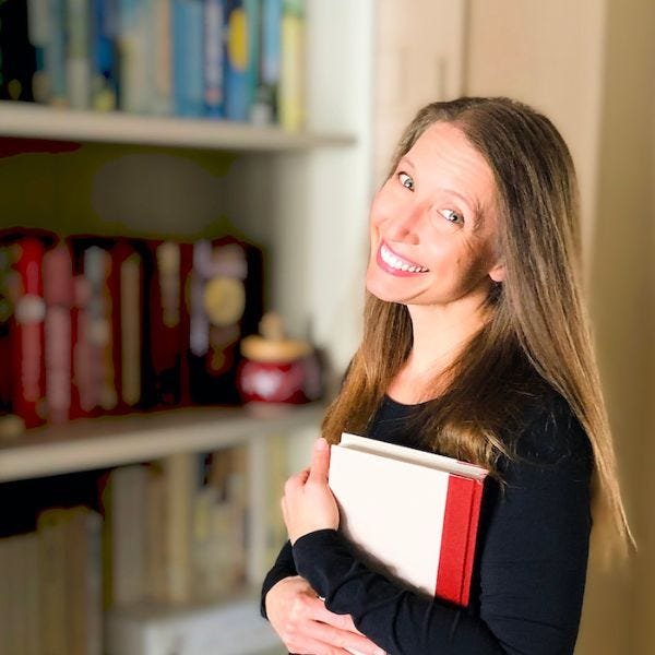 Image of writer Michelle Watson of Library Binding standing in front of a shelf full of books, holding a book in one arm and smiling at the camera